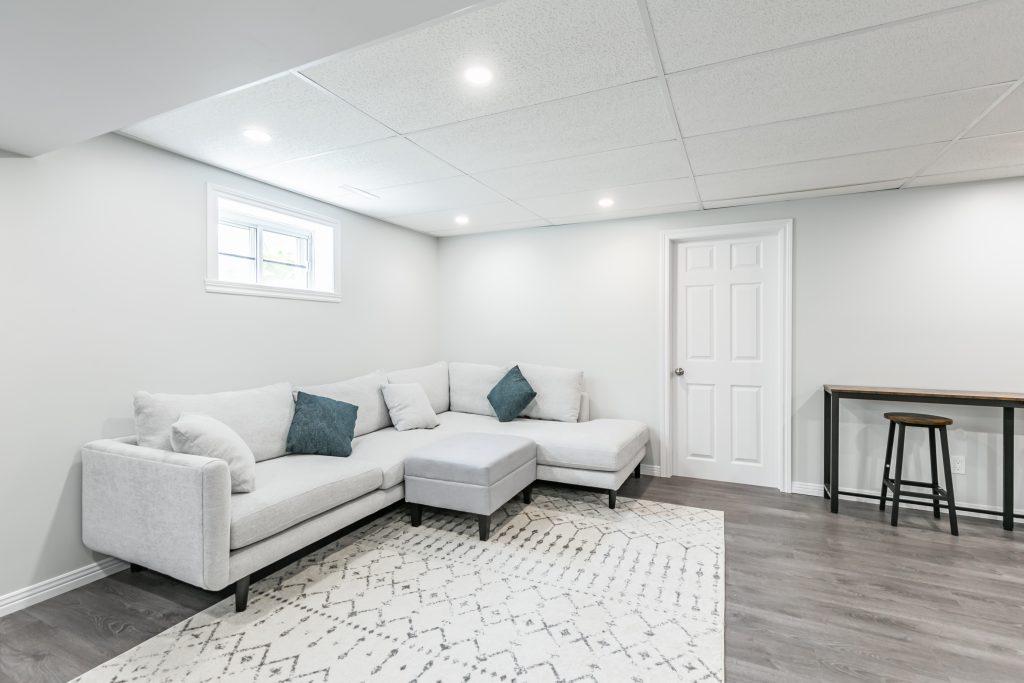 Sous-sol à murs blancs, décoré d’un canapé gris pâle modulaire, avec plancher en bois et tapis à motifs géométriques