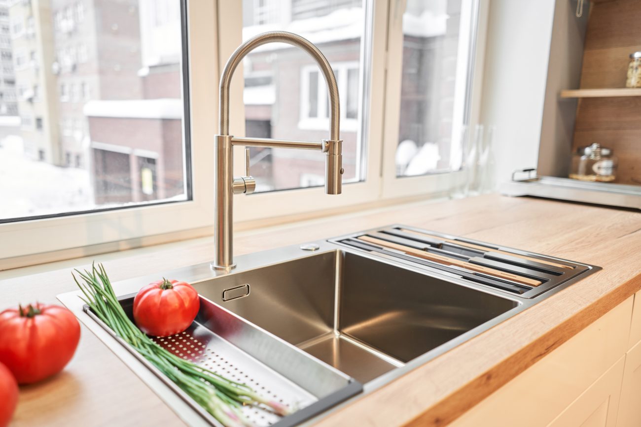 Évier en inox devant une fenêtre, tomates et oignons verts sur un comptoir de cuisine en bois