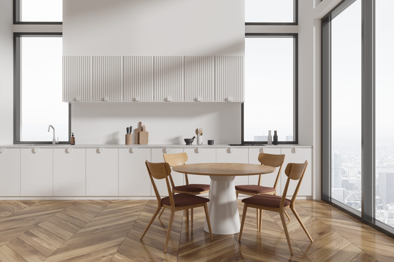 Modern kitchen in apartment with white walls, wooden floor, white cupboards and cabinets and round dining table with chairs