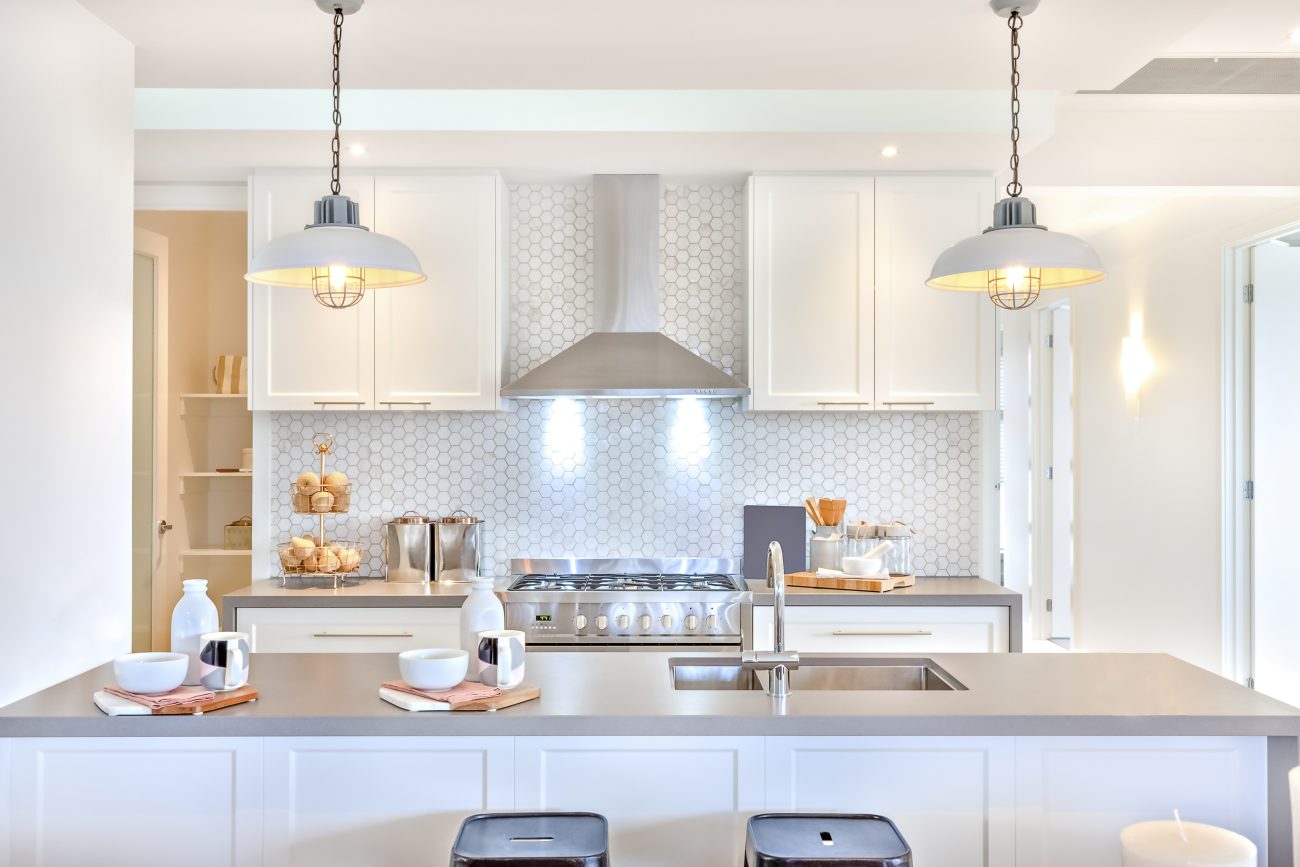 In a modern white and pale grey kitchen, backsplash of hexagonal tiles, suspended industrial lamps and large island with sink