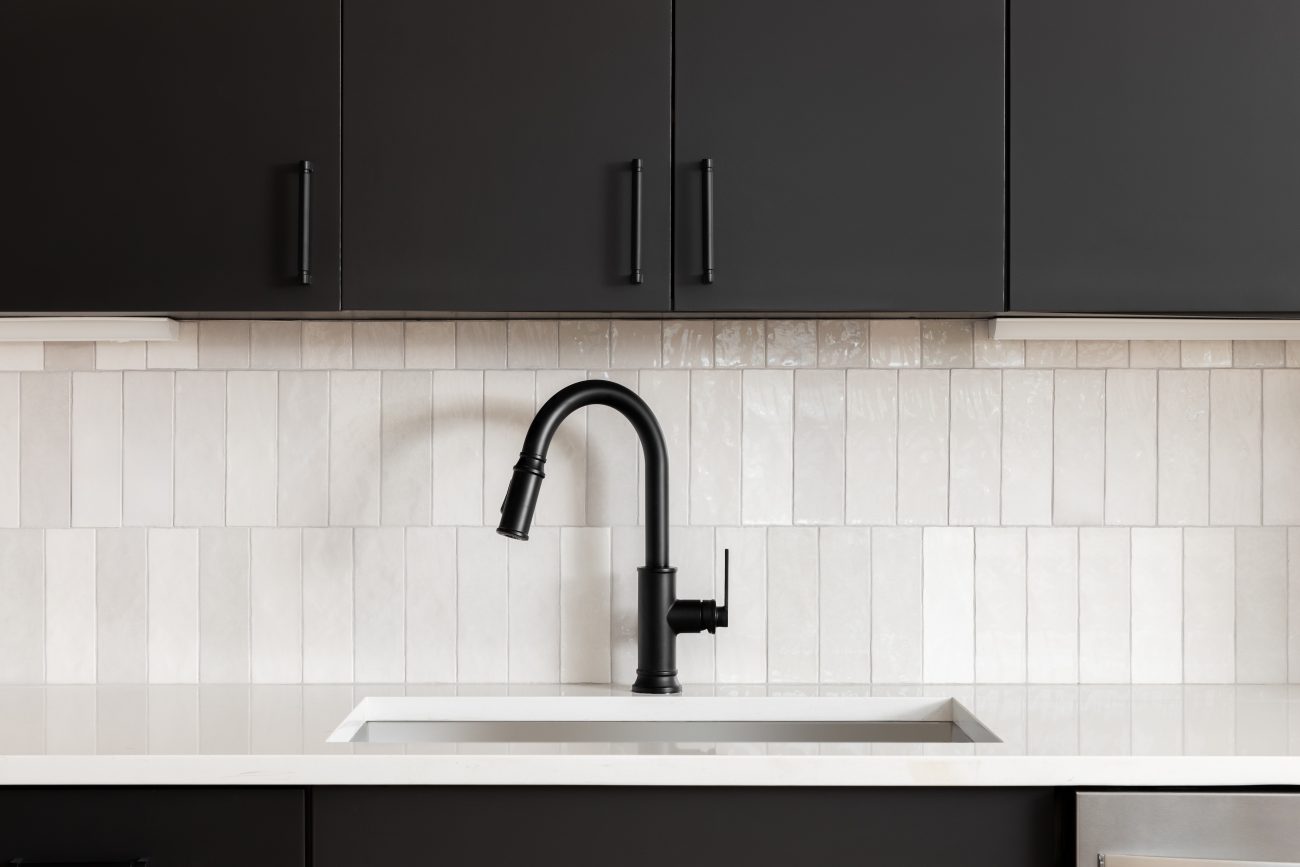 In a modern kitchen, black cabinets, vertical subway tile backsplash, black faucet and white marble countertop