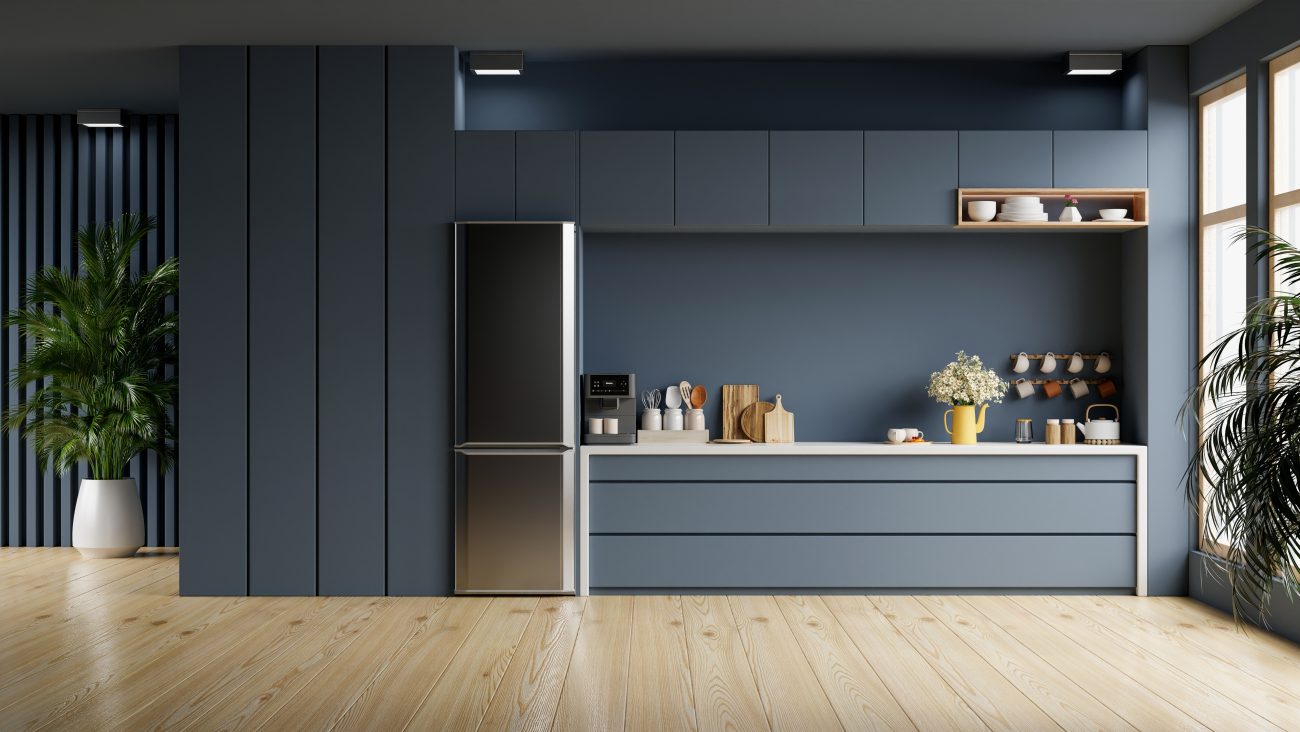 Cupboards and fridge in the alcove of a dark grey kitchen, countertop accessories including coffee pot, wooden chopping board, flowerpot and mugs