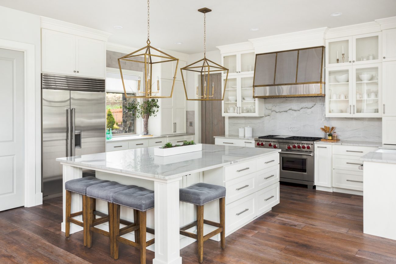 White modern kitchen with grey velvet upholstered stools, large island with marble countertop, large refrigerator and glass cabinets