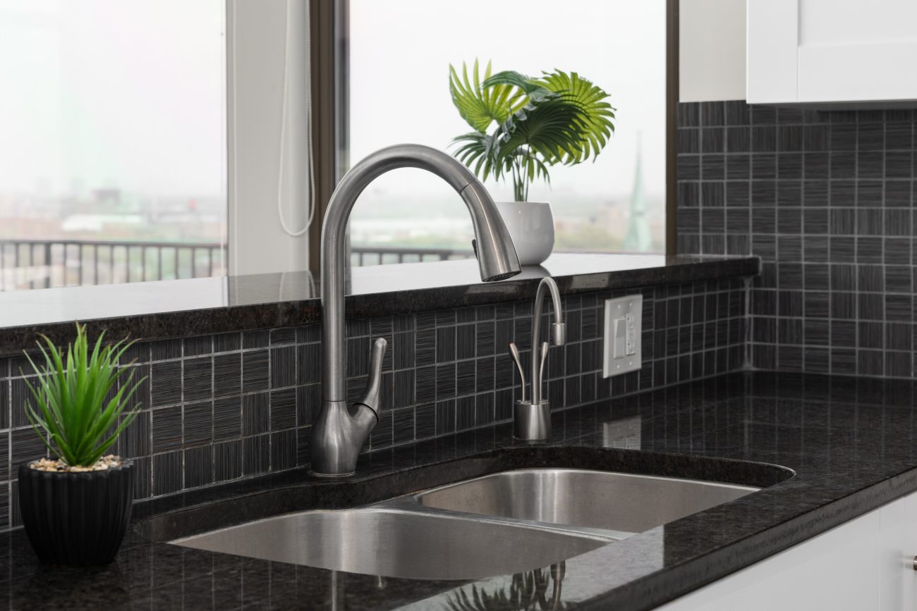 In a modern kitchen, sink detail with a square dark grey tile backsplash, black granite countertop, and a brushed chrome faucet