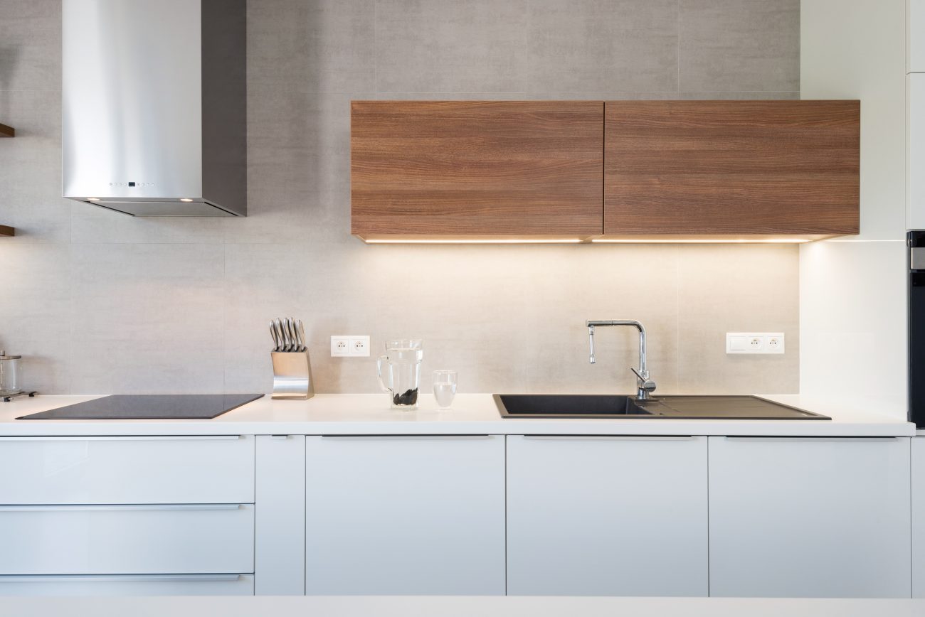 In a modern white kitchen, stainless steel cooker hood, wood cabinets with indirect lighting and black sink