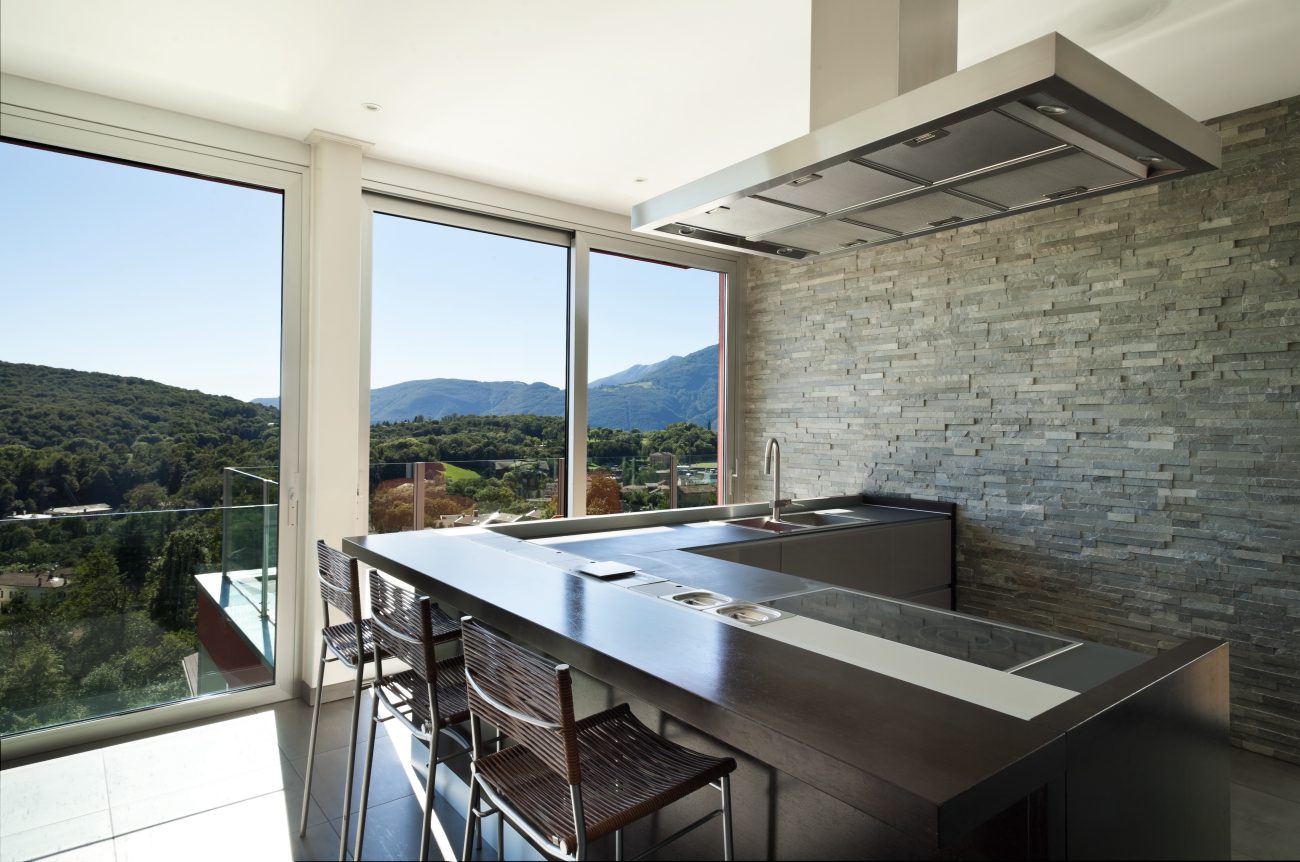 Picture windows with mountain views in a modern kitchen with gray brick wall and L-shaped island