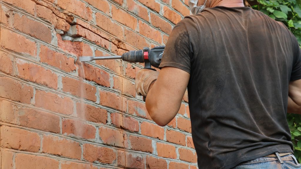 Worker holds a drill to remove the mortar from a red brick wall