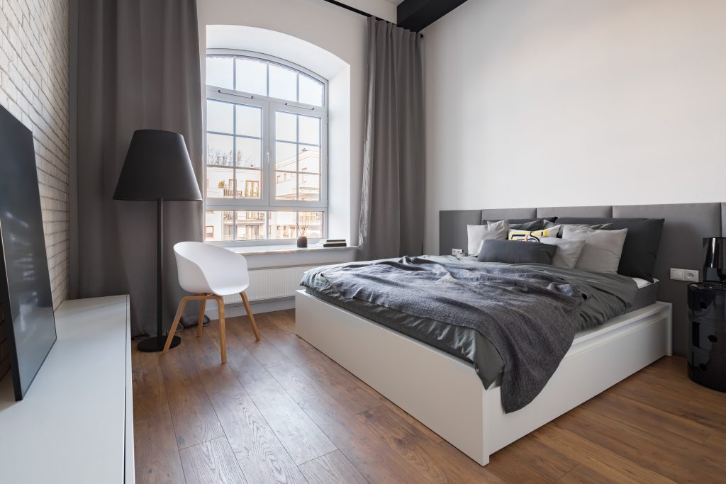 Chambre à coucher à plafond haut, mur de briques blanches, lit à couette grise, rideaux gris et grande fenêtre à carreaux et haut en arche, avec vue sur multiplex