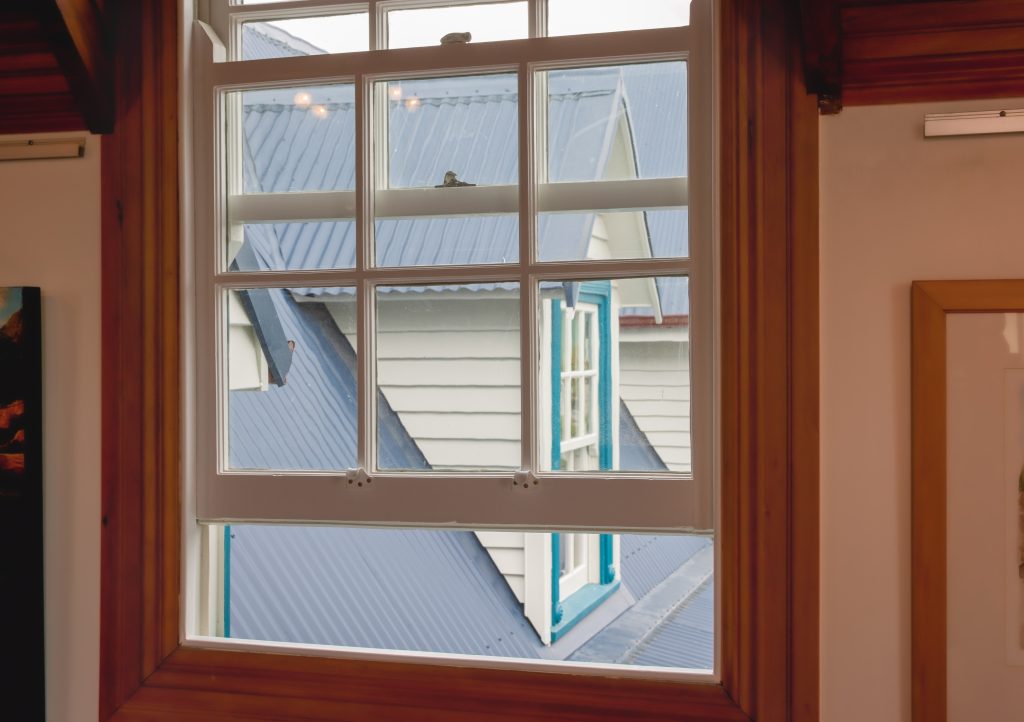 Double-hung window open, with white mullions and a brown wooden frame, overlooking sloping roofs and the gable of a house