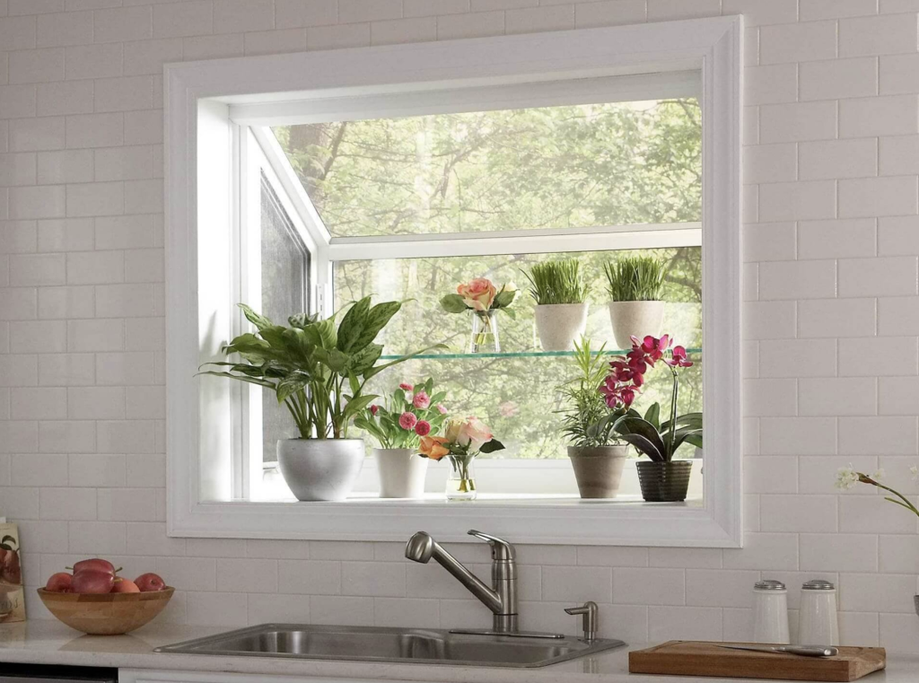 In a kitchen with a white ceramic tiled wall, garden style window above the sink, with a glass shelf decorated with plants and flowers