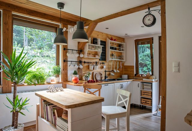 White farmhouse kitchen with wall, counter, butcher block, wooden chairs and beams, industrial light fixtures, retro clock