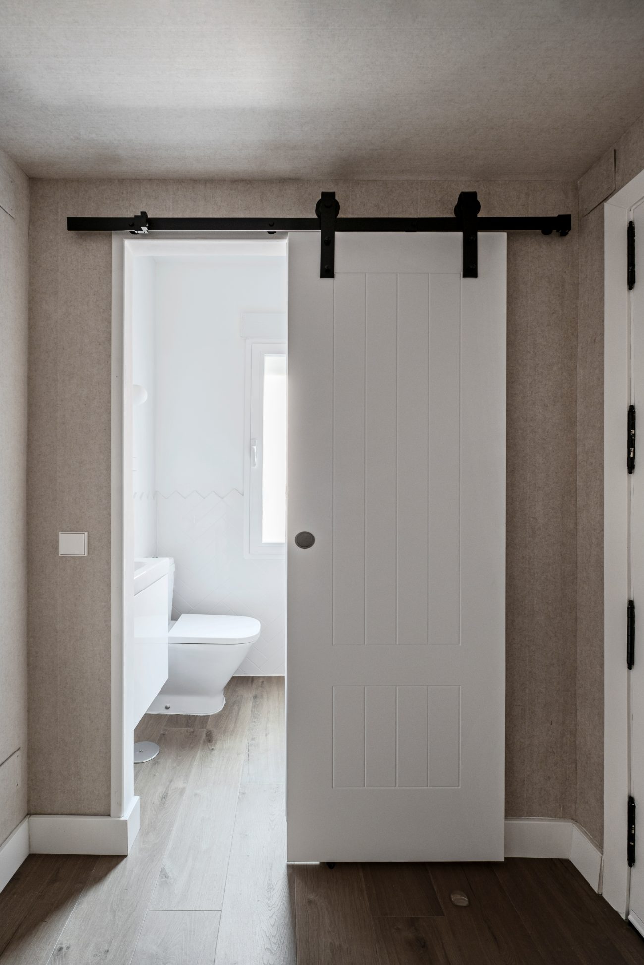 White barn door on a black rail, slightly open to a small white bathroom with a toilet and vanity