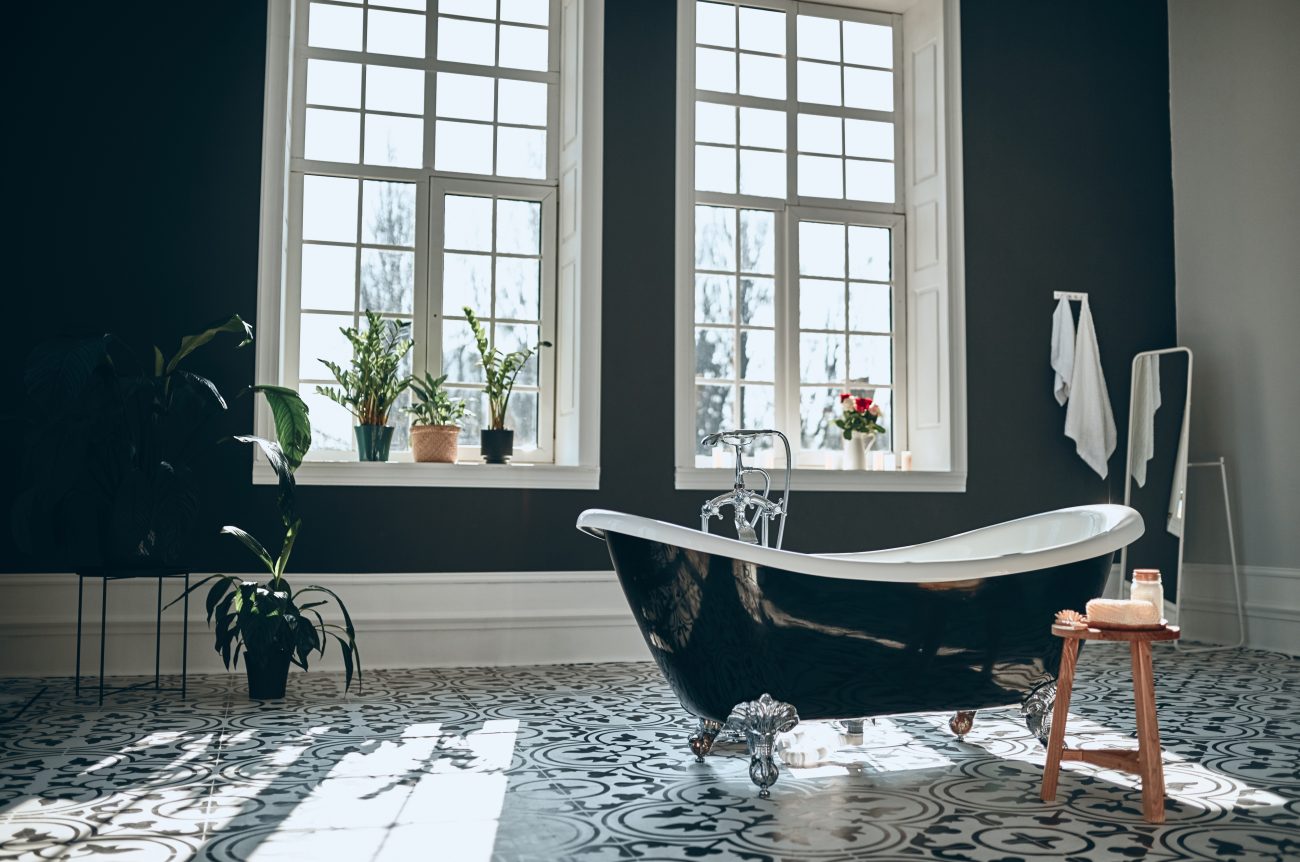 Vintage-style freestanding black bathtub with white interior and silver feet in a luxurious setting: black wall, large windows with white mullions, several plants, abstract patterned floor, small wooden bench with beauty products