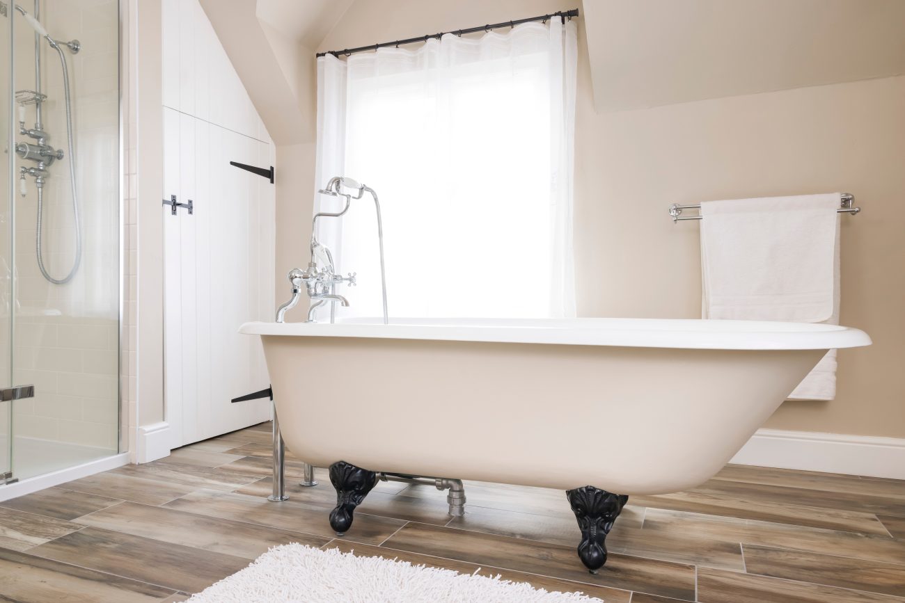 Freestanding white bathtub with black legs, in a sand-coloured bathroom, barn door, white voile curtain in the window, and wood plank floor