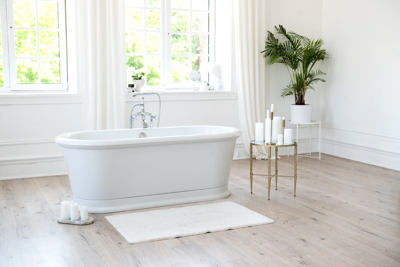 In an all-white bathroom with windows overlooking a green landscape, a freestanding oval bathtub with silver hardware, candles placed on the light wood floor and on a side shelf