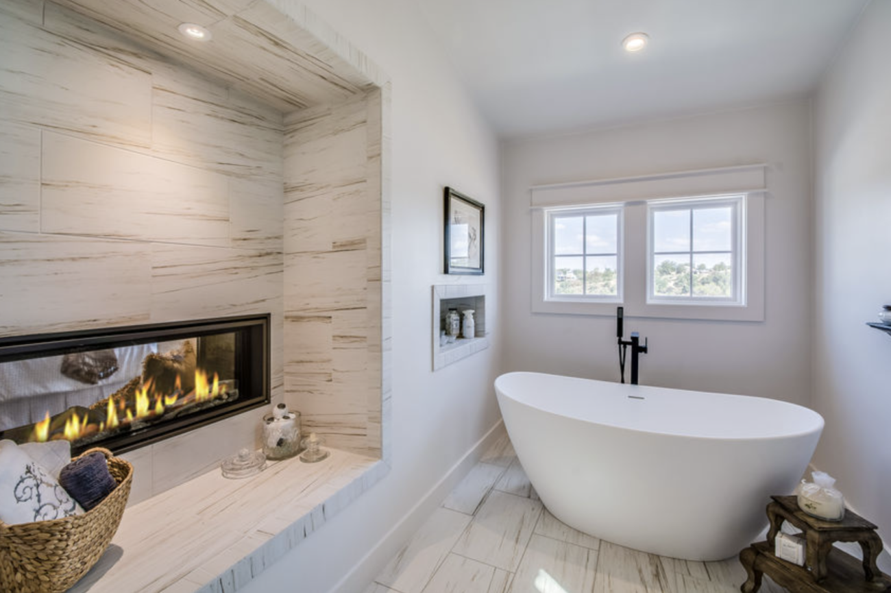 Luxury bathroom with a very wide gas fireplace built into an alcove containing a woven basket, towels, and bottles, a white oval bathtub, and two paned windows