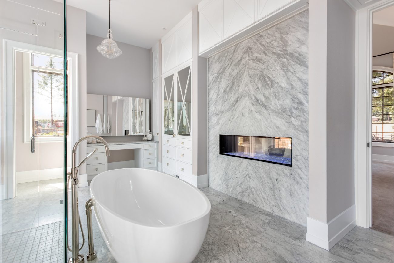 In a pale grey and white luxury bathroom, a freestanding bathtub with a sliding bar shower, a two-sided gas fireplace built into a veined marble partition, a makeup corner with a vanity and mirror