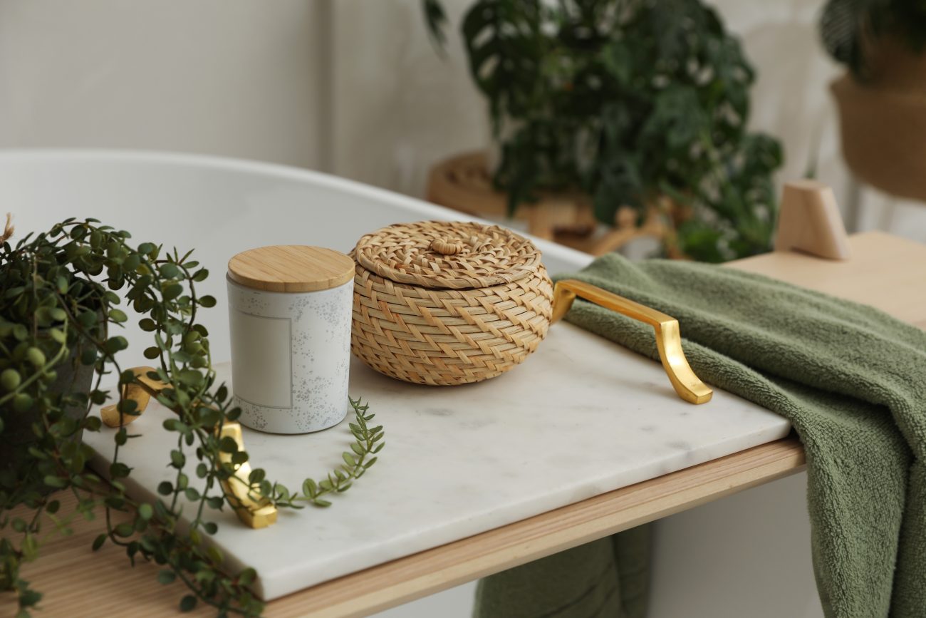 On a freestanding white bathtub, a wooden tray and a marble tray with gold handles, holding a candle, a small woven basket, a plant, and an olive-colored towel