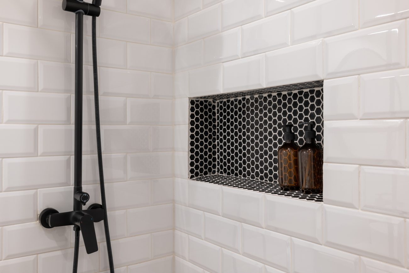 Shower with glossy white subway tile walls, black hardware, wall niche with black and white hexagonal mini tiles, and two care product bottles