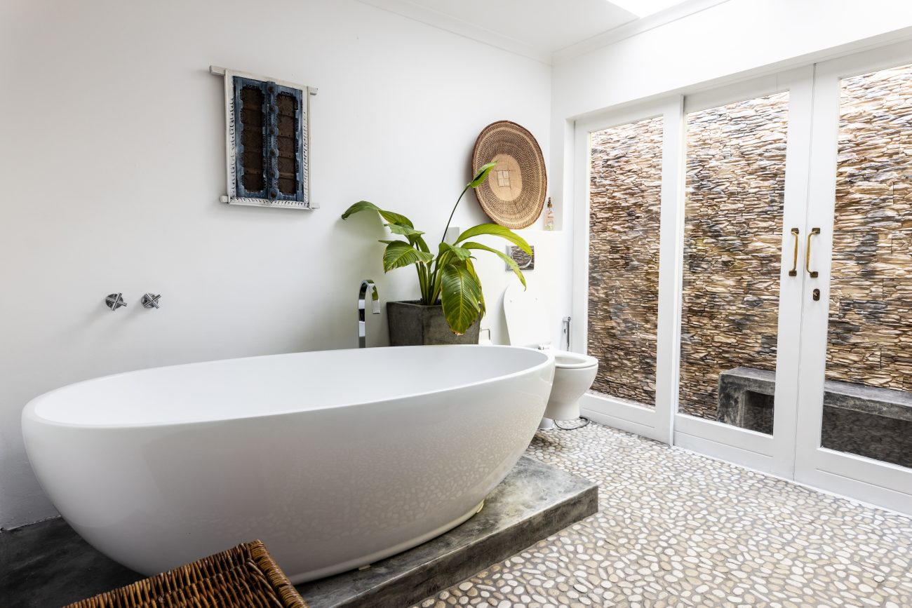 Freestanding oval bathtub on a marble platform, abstract canvas mounted on a wooden frame, woven basket hanging on the wall, large-leafed plant, pebble floor, and sliding doors opening onto a layered stone wall