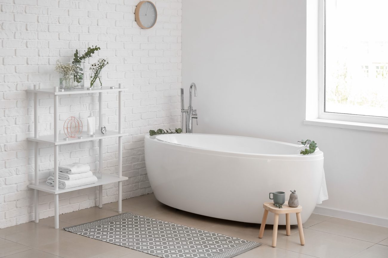 Freestanding oval bathtub in an all-white minimalist decor with a brick wall, a shelf unit for towels, eucalyptus branches in pots and on the edge of the tub, a small wooden bench, and a rug