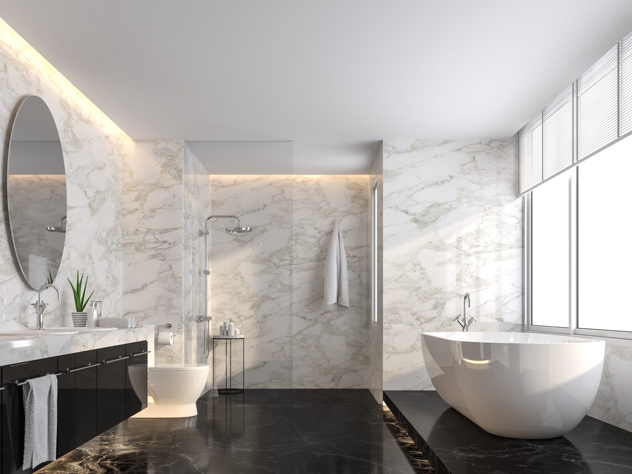 Very bright luxurious bathroom with walls covered in white marble with grey veins. The white porcelain bathtub and toilet contrast with the black floor and vanity. A glass partition separates the walk-in shower at the back of the room