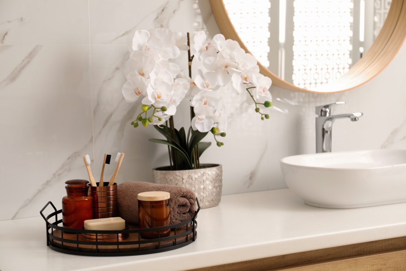 In a luxury bathroom, a vanity with an oval basin, a tray with a candle, toothbrushes, soap bars, a white orchid, marble-covered wall, and round mirror with a wooden frame