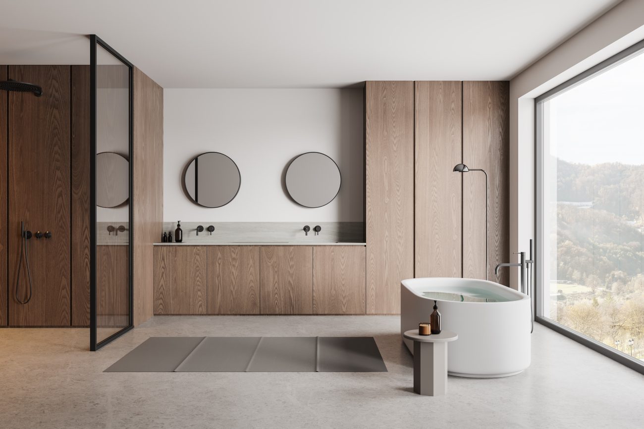 In a minimalist luxury bathroom, handleless ribbed wood storage cabinets, two sinks topped with two round mirrors, a freestanding white bathtub, and a bay window with a mountain view