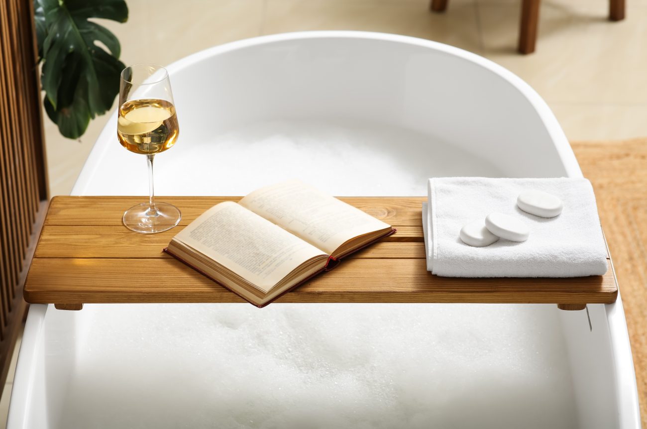 On a freestanding white bathtub filled with bubble bath, a wooden tray holding a glass of white wine, an open book, a folded white towel, and soap bars