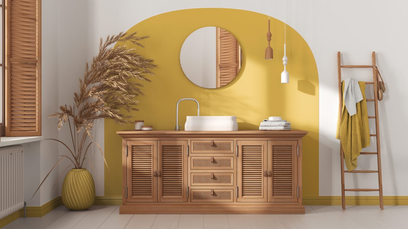 In a luxurious room, a wooden storage unit with louvred doors and cane drawers, a white basin, a round mirror on a yellow accent wall, and a ladder serving as a towel rack