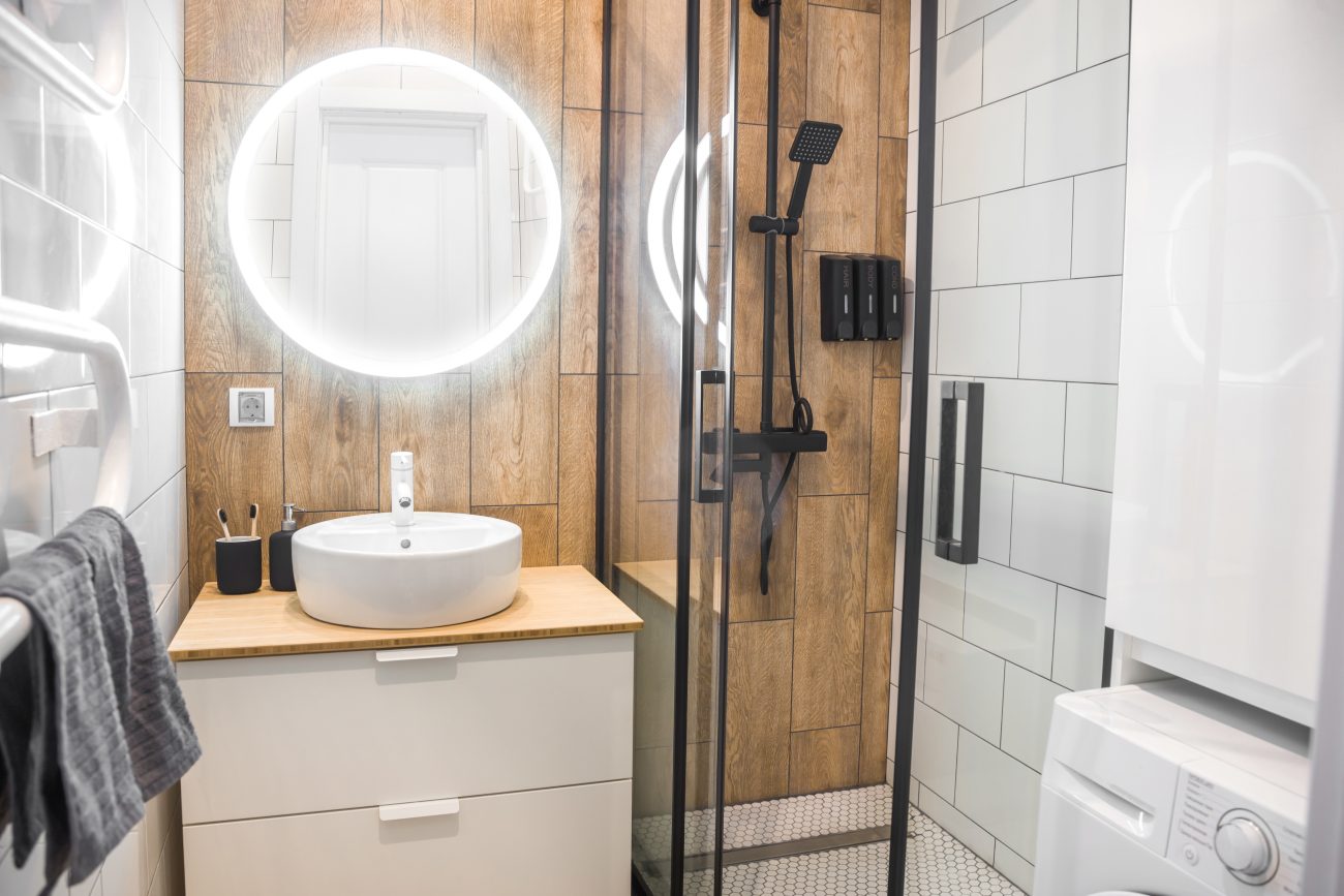 Small bathroom with wood-effect tiles and white subway tiles on the walls, featuring a corner shower with a black frame, two-drawer vanity topped with a round basin, backlit round mirror, and washer