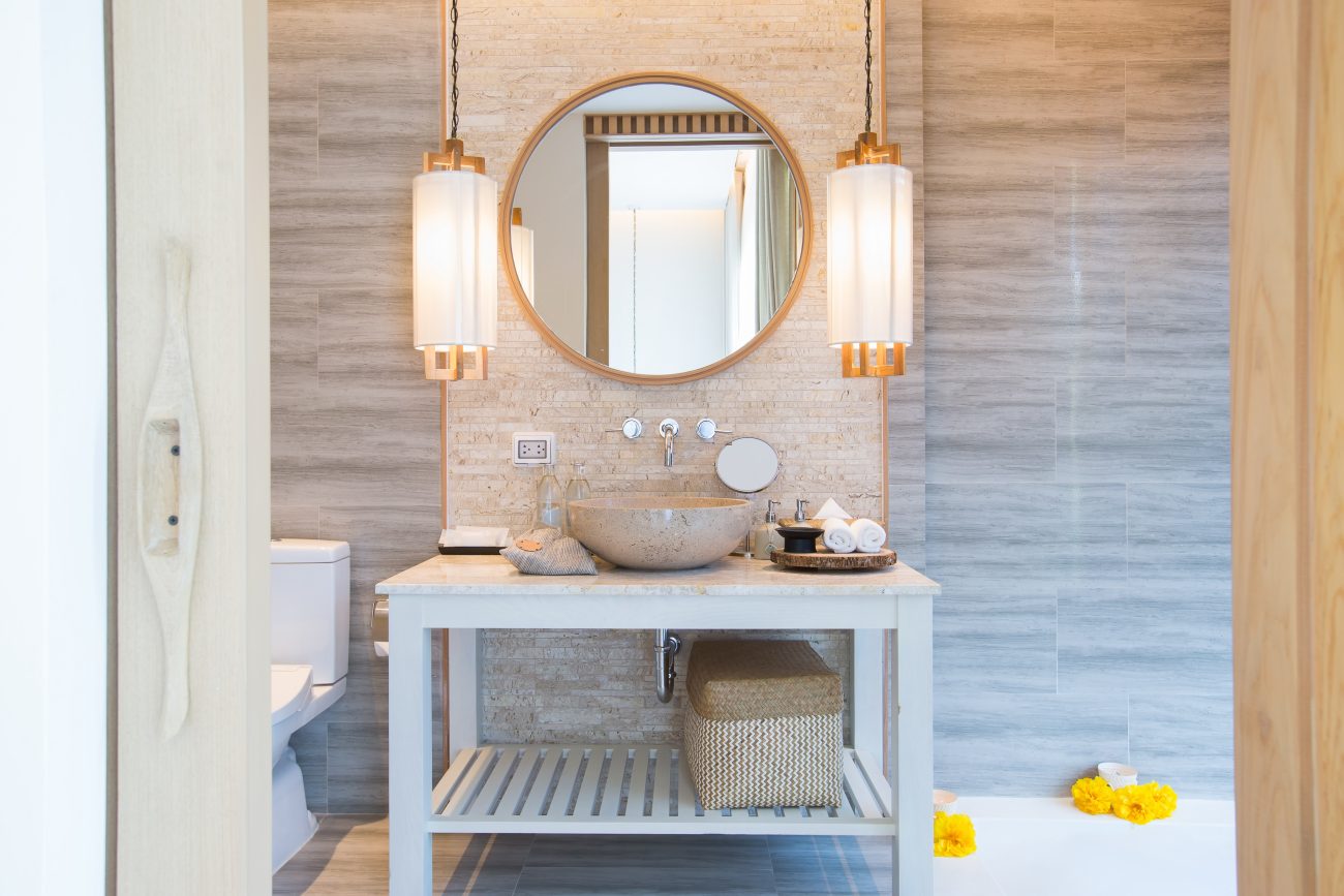 Dans une salle de bain de luxe, une table à comptoir en marbre sur lequel sont déposés une vasque en grès beige, des serviettes, des flacons de produits de soin. De chaque côté du miroir rond, une suspension de type lanterne japonaise, avec structure en bois et abat-jour blanc diaphane