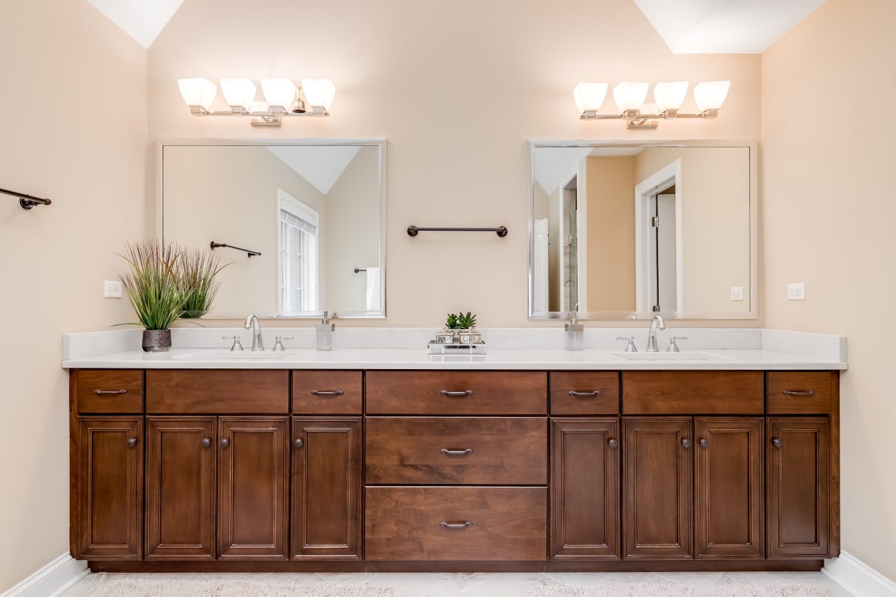 Dans une salle de bain bien éclairée par des appliques murales, un meuble de rangement très large de style Shaker, deux miroirs et une plante dans un coin