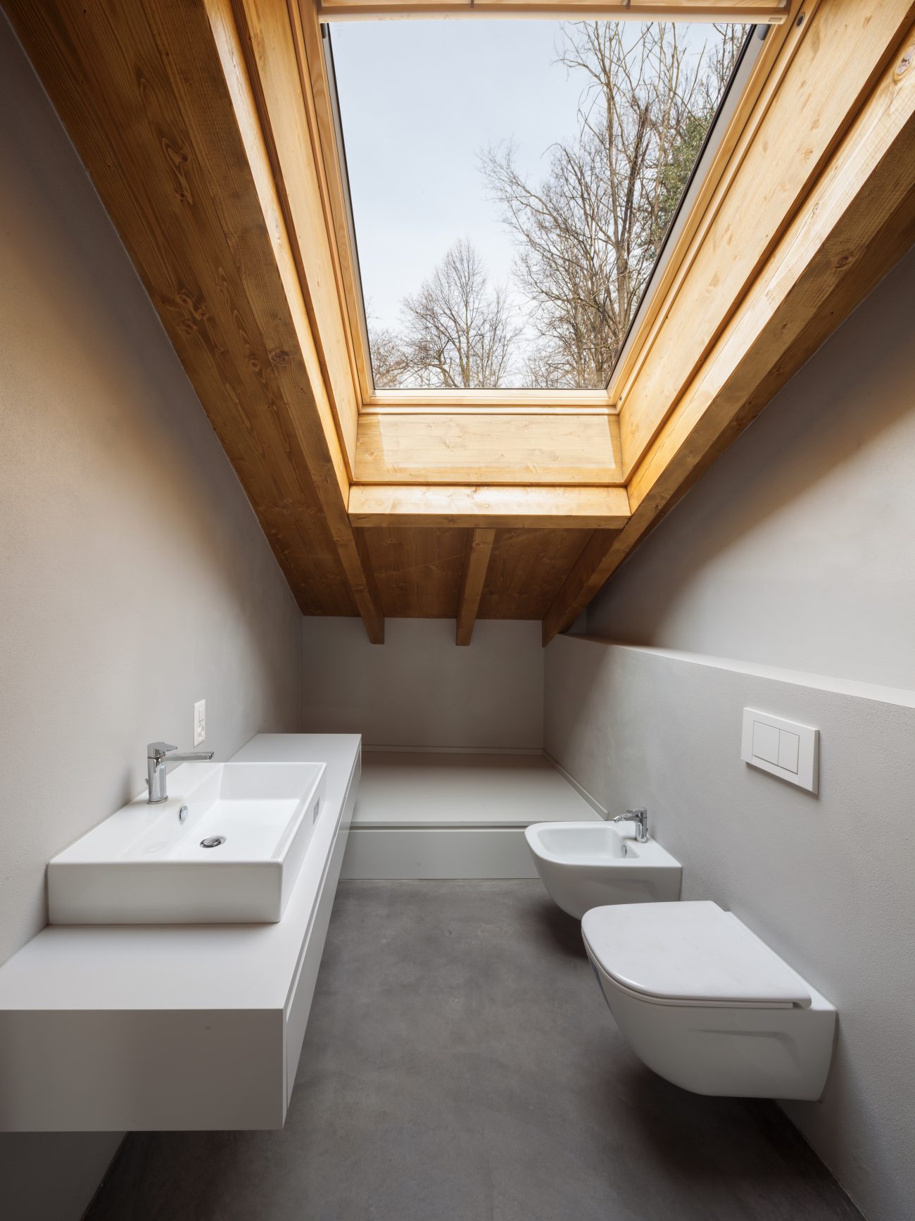 Salle de bain grise aménagée sous les combles, avec sanitaires, meuble-lavabo et vasque blancs, puits de lumière en pente monté sur cadre de bois