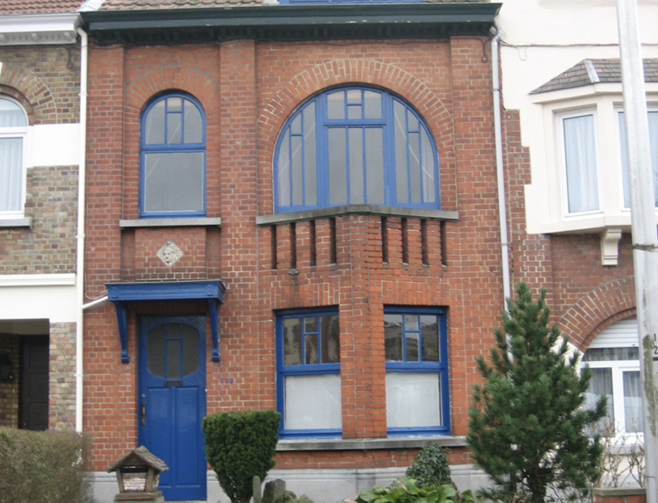 Édifice à deux étages en briques rouges abimées, avec fenêtres en demi-lunes et porte bleue