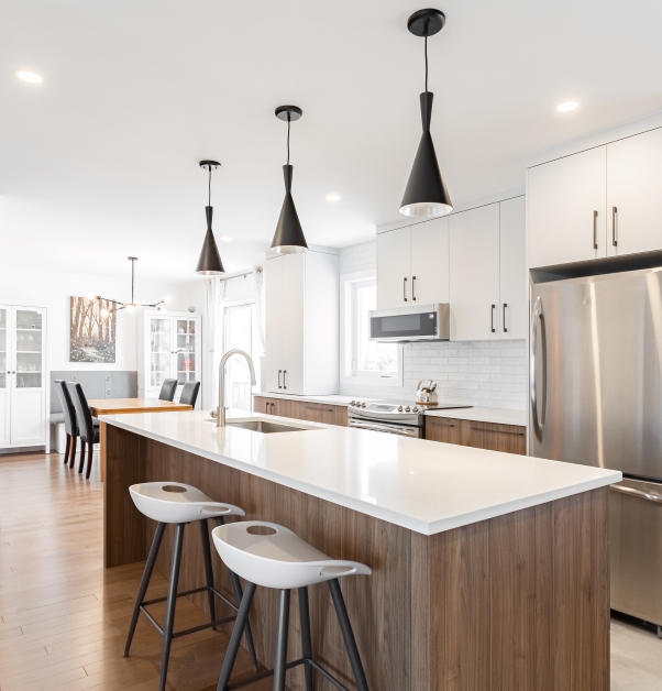 Modern white kitchen with white cabinets, large laminate island with white stone countertop, black pendant lighting, white ceramic floor and natural toned hardwood floor.