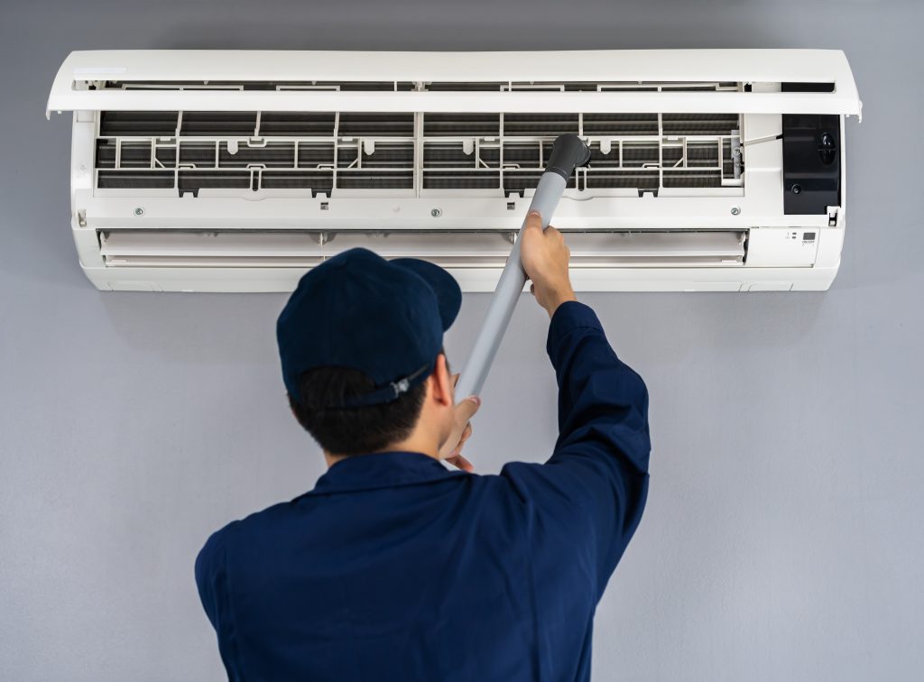 Technicien en uniforme et casquette bleu marine, utilisant un aspirateur pour nettoyer le climatiseur mural