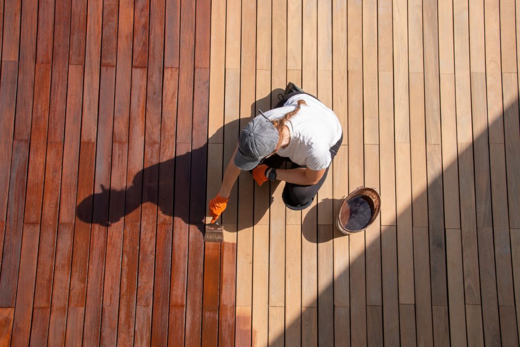 Femme avec une longue tresse, une casquette blanche et des gants orange, entretient une terrasse en bois avec de l’huile pigmentée