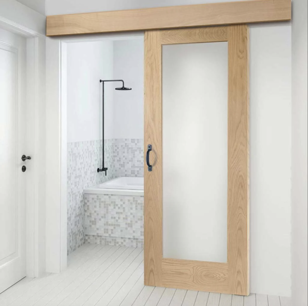 Dans une salle de bain, porte coulissante vitrée à cadre de bois naturel qui sépare l’espace réservé à la baignoire, mur et côté de la baignoire couverts de minituiles grises et blanches. 