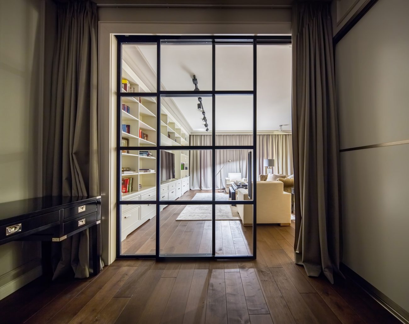 Black slatted sliding French doors framed by curtains, opening onto a living room adorned with a large wall unit