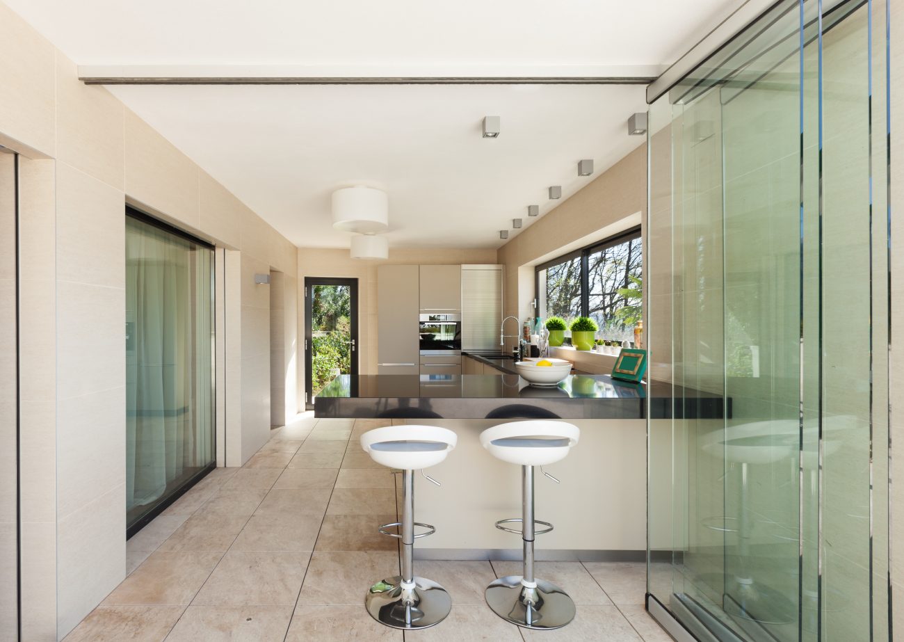 Glass folding doors opened onto a narrow kitchen with an L-shaped black countertop and two white stools with chrome column bases