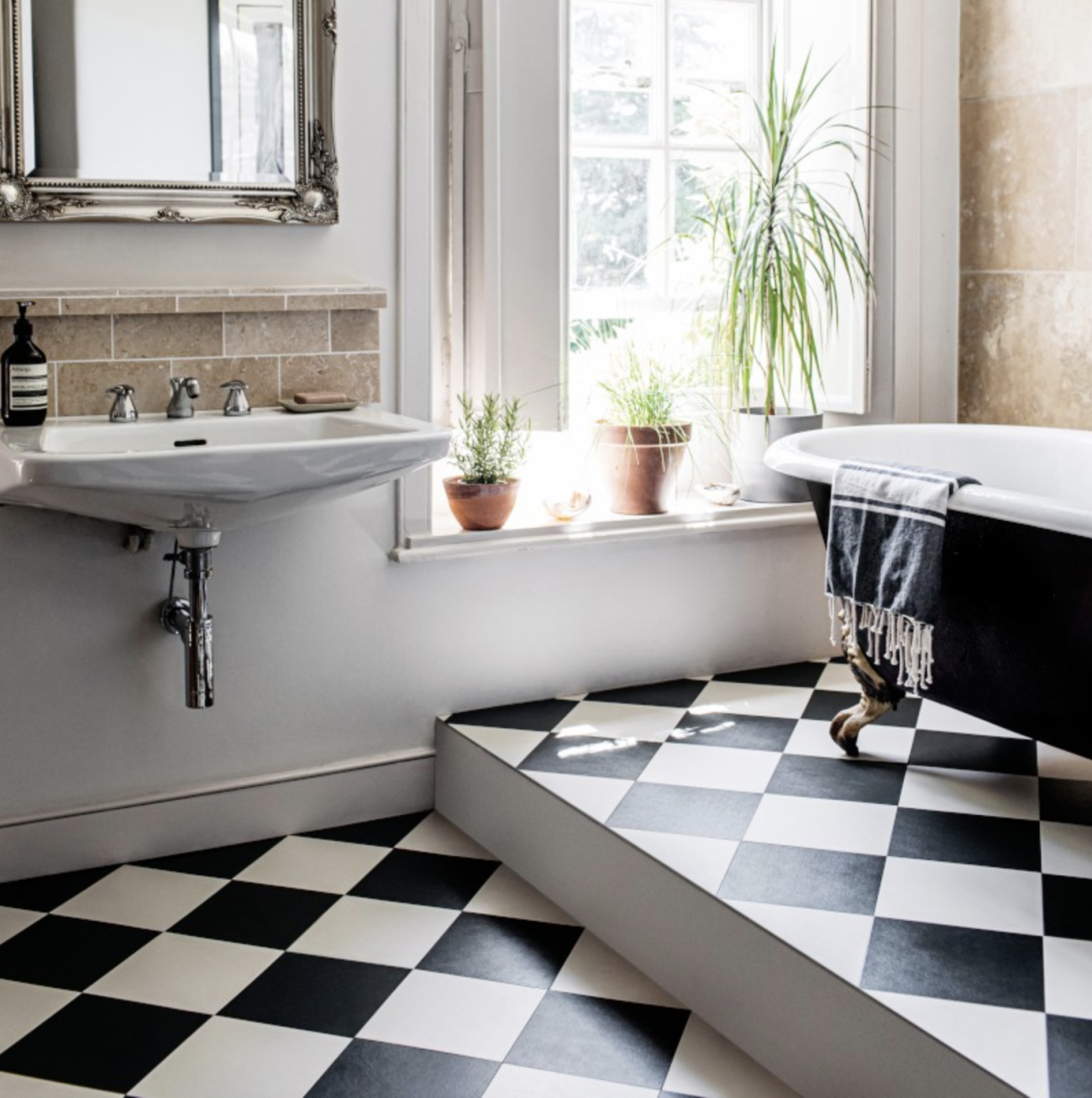 Retro bathroom with black and white vinyl tile floor, black clawfoot bathtub, travertine-adorned wall, retro wall-mounted ceramic sink, and stylized frame mirror