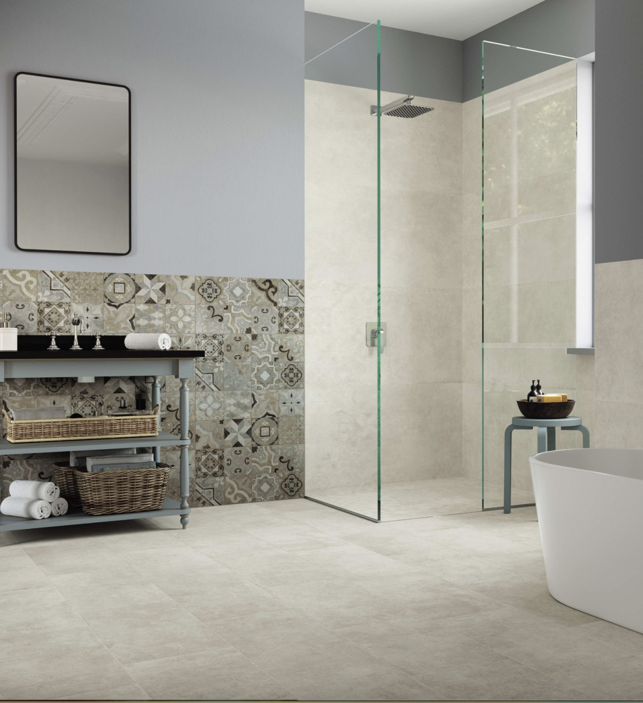 A spacious bathroom in shades of grey and beige, featuring a glass-walled shower, an oval bathtub, a small bench, and a grey-painted wooden vanity with rattan baskets. The entire shower is clad in marble-imitating cement tiles, and a half-wall is decorated with a mosaic of tiles with various geometric patterns and flourishes