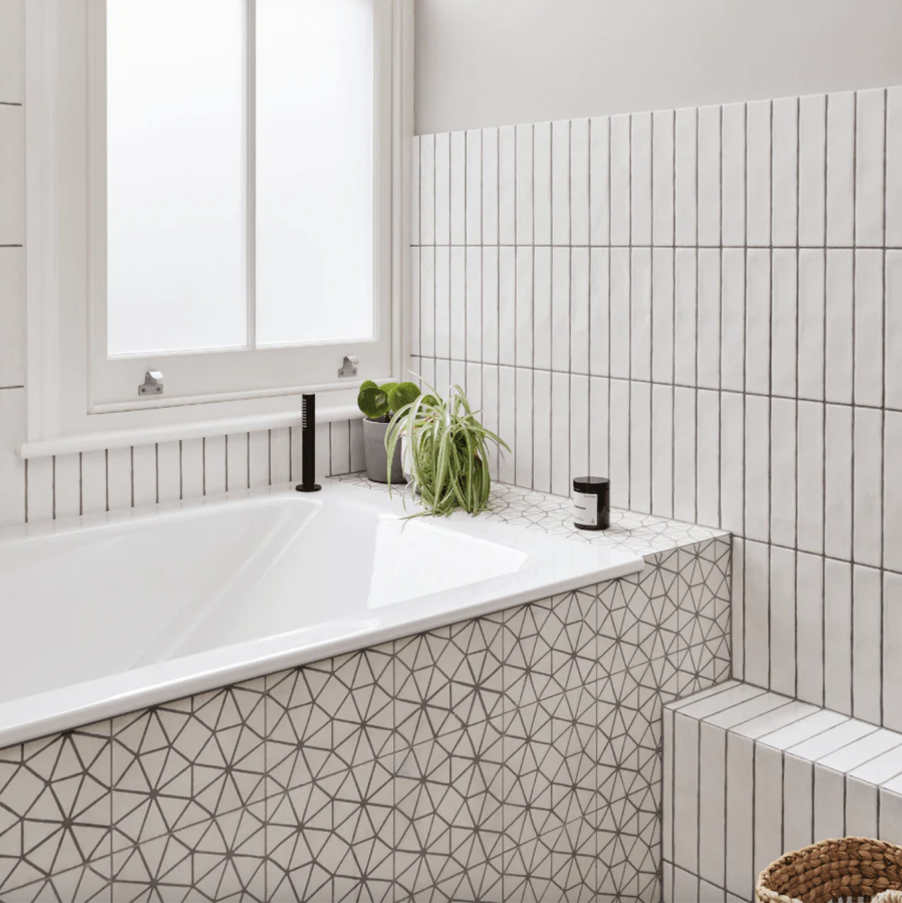 In a white bathroom, an embedded rectangular bathtub, wall covered with white cement tiles featuring kaleidoscope designs, half-wall decorated with vertical white tiles two green plants in the corner