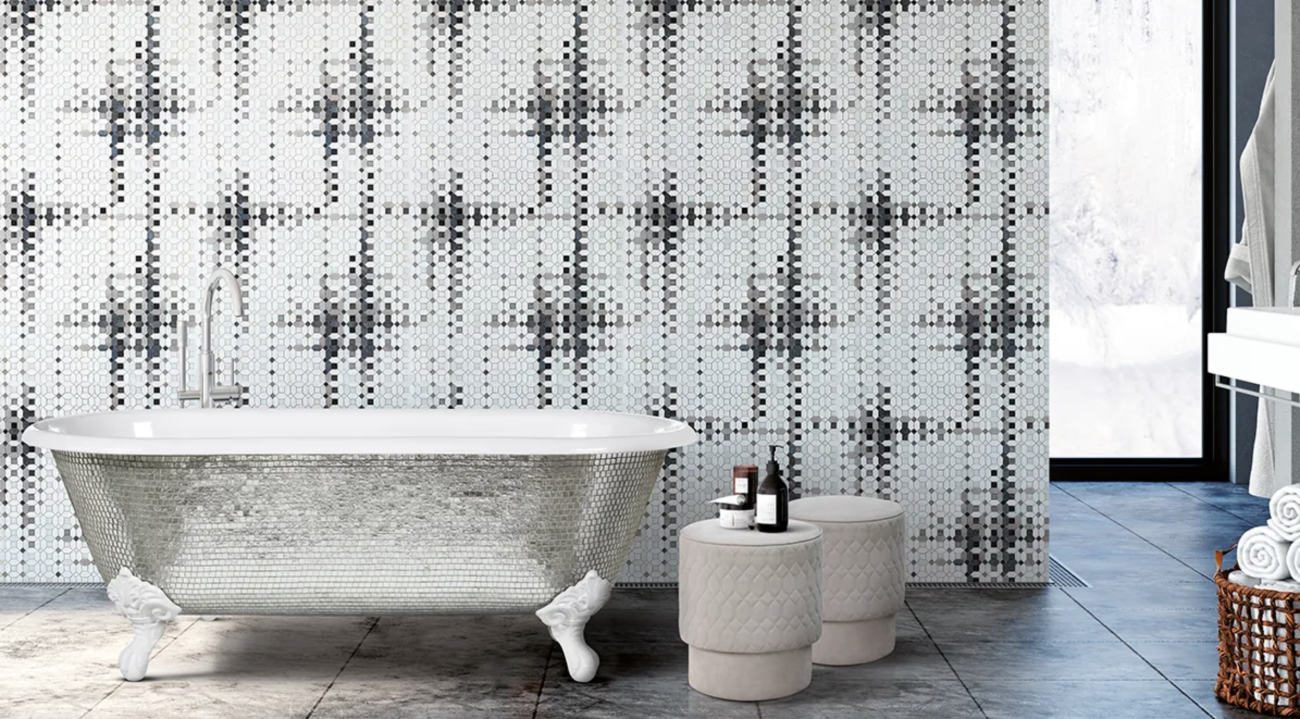 Bathroom with clawfoot bathtub decorated with silver mini-tiles, wall covered with white, grey, and black mosaic, velvet poufs used as tables for beauty products, and woven basket with rolled towels