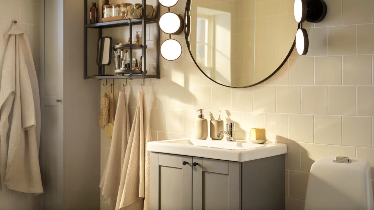 In a bathroom, butter yellow tiled wall, taupe two-door vanity unit with surface-mounted basin, circular mirror surrounded by flat spotlights, shelves filled with various beauty accessories