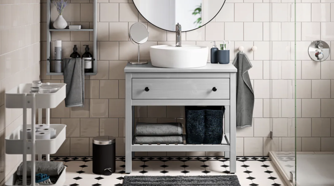 Bathroom with walls covered in greige tiles and cabochon floor tiles in cream and black, open shelving filled with bottles, ceramic basin on a storage unit, table mirror, towels, and woven basket