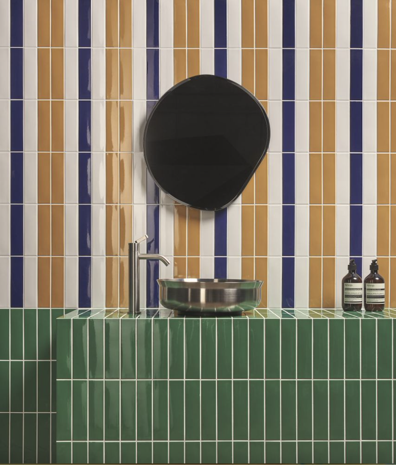 Bathroom wall decorated with vertically aligned rectangular tiles in ochre, indigo, and cream white porcelain, organic-shaped mirror, stainless steel basin on a full-height half-wall clad in basil green porcelain tiles