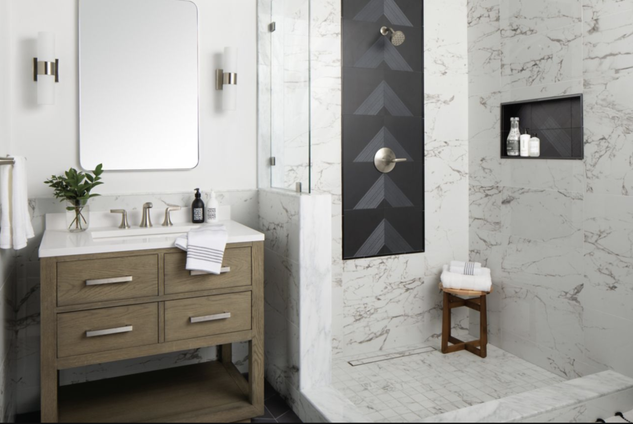 In a bathroom, open shower with white marble and grey veining, niche for beauty products, black panel on one wall, and small wooden bench. Four-drawer vanity, oval-cornered mirror, and wall sconces