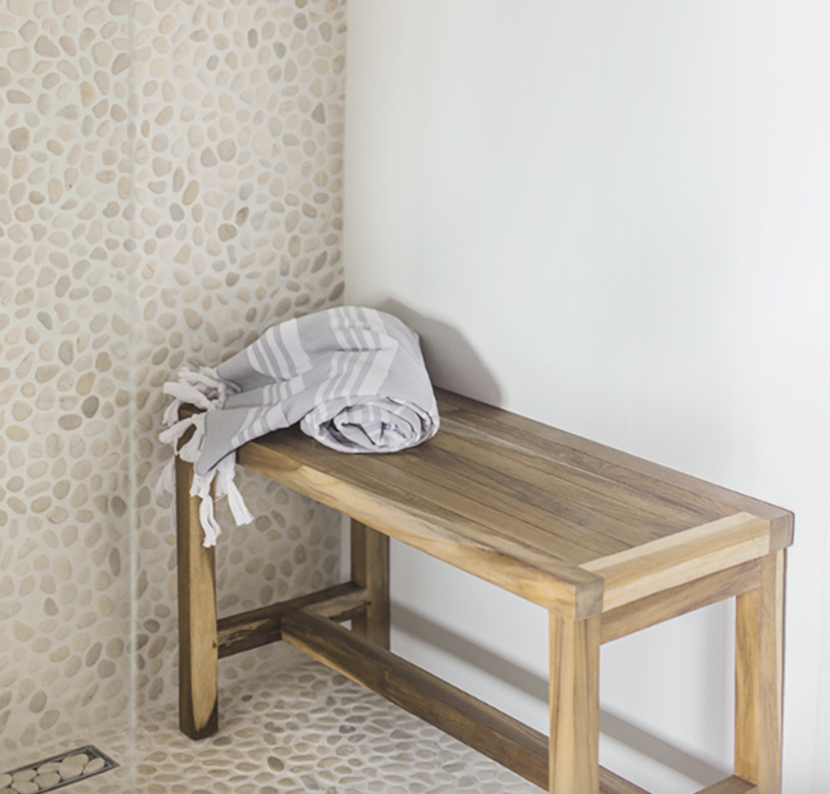 In a bathroom, shower with walls and floor covered in pebbles, niche for hair products, wooden bench, and fouta